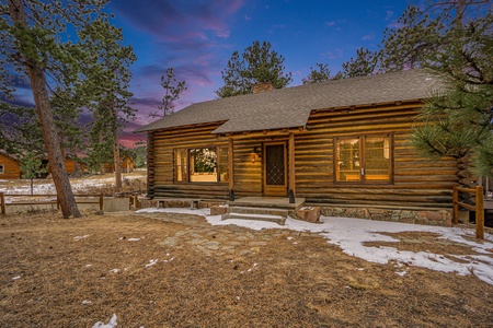 Log cabin surrounded by trees with light snow on the ground, set against a colorful twilight sky.