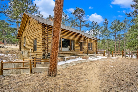 A rustic log cabin surrounded by pine trees on a clear day, with a visible dirt path and wooden fencing.