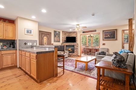 Open-concept living space with a kitchen, stone fireplace, TV, wooden floors, and a dining area. Cozy seating with a bear sculpture on a side table. Natural light from large windows.