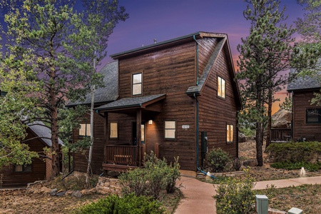 A wooden cabin with a porch is surrounded by trees. The sky is a vibrant mix of purple and orange hues, suggesting sunset. A walkway leads to the entrance.