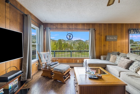 Living room with a sectional couch, rocking chair and ottoman, flat screen TV and a large window with beautiful mountain views