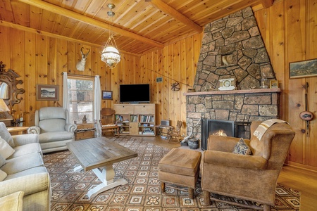 A cozy living room with wood-paneled walls, a stone fireplace, two armchairs, a couch, a wooden coffee table, a patterned rug, and a mounted TV.