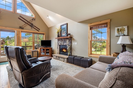 Living room with fireplace, 2 leather arm chairs, small round table, sofa that seats 4, 3 small ottomans, flat screen tv, and windows with mountain views.