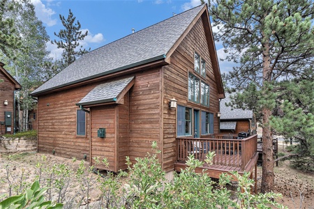 Wooden cabin with a large deck set among pine trees, featuring a sloped roof and multiple windows.