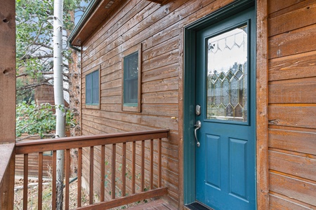 Wooden cabin exterior with a blue door and two windows. A railing lines the small porch area, and trees are visible in the background.