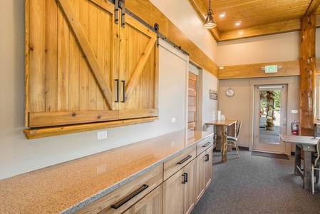 Kitchen area with wooden cabinets, a sliding barn door, a countertop, and tables. A clock and exit sign are visible. The space has a high ceiling with wood accents.