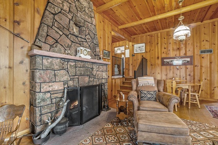 A rustic living room features a stone fireplace, wooden walls, an armchair with ottoman, a wooden chair, a small dining table, and a chandelier hanging from the ceiling.