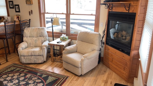 Two armchairs near a large window and a fireplace in a cozy living room with a wooden floor and a patterned rug.