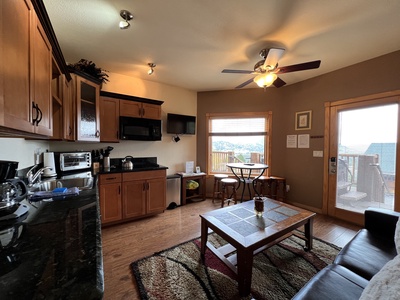 A cozy kitchen and living area with wooden cabinets, a small dining table, a ceiling fan, and a sliding glass door opening to a deck with a view.