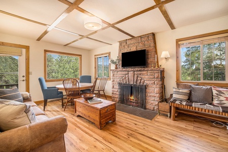 Kittery West Estes Park, Living room with sofa that seats 3 bench coffee table, bench seat with seating for 2, flat screen tv, wood burning fireplace, round dining table with seating for 5, window with view of mountains, and door leading to deck.