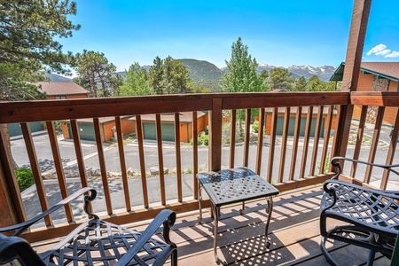 A wooden balcony with two chairs and a small table overlooks a residential area with trees and mountains in the background on a clear day.