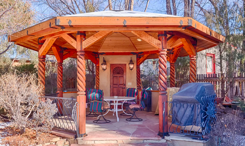 A wooden gazebo in a backyard with a grill.