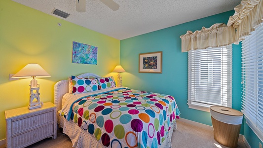 A brightly decorated bedroom with yellow and teal walls, a bed with a colorful polka dot comforter, two bedside lamps, a painting, a window with white blinds, and a wicker nightstand.