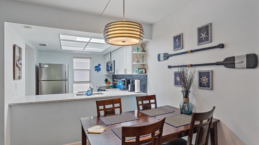 A small, modern kitchen and dining area with a wood table, four chairs, wall decor of paddles, and nautical-themed art. The kitchen features a stainless steel refrigerator and an open shelf design.