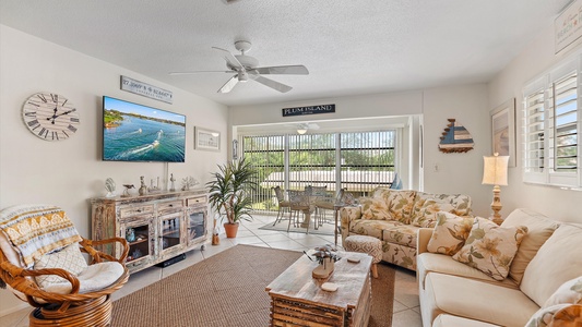 A living room with a coastal theme, featuring a sofa, a wicker chair, a wooden coffee table, a TV, a ceiling fan, and decor. A sliding door opens to a sunroom with a table and chairs.