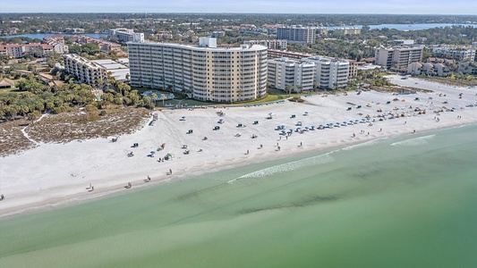 Aerial view of a coastal city featuring a wide sandy beach, ocean waves, beach chairs, umbrellas, and several mid-rise buildings and resorts.