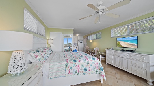 Bright and cozy bedroom with a floral bedspread, a ceiling fan, TV, and white furniture; window showing beach view on the TV screen.