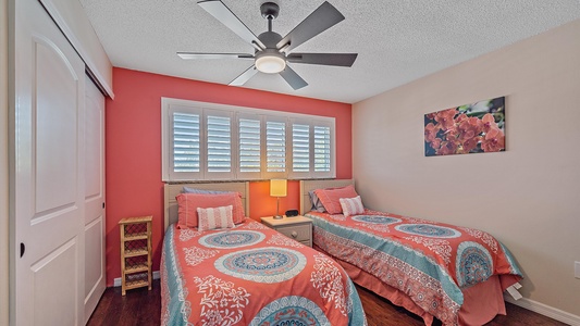 A bedroom with two twin beds featuring pink and blue bedding, a nightstand with a lamp between them, a ceiling fan, a window with shutters, a painting on the wall, and a sliding door closet.