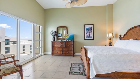 A bright bedroom with a large window, a wooden bed, a dresser with a mirror, a small TV, and a chair. The room has tiled flooring and a view of the ocean through the window.