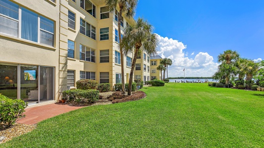 A multi-story beige building with numerous windows and balconies is shown next to a well-maintained grassy lawn with palm trees, leading toward a body of water and a blue sky with scattered clouds.