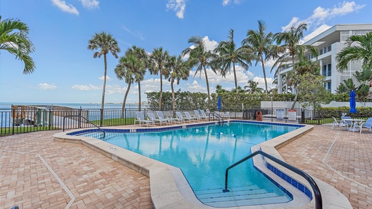 An outdoor pool area with lounge chairs, palm trees, and a view of the ocean. A modern building is visible on the right side. The sky is clear with a few clouds.