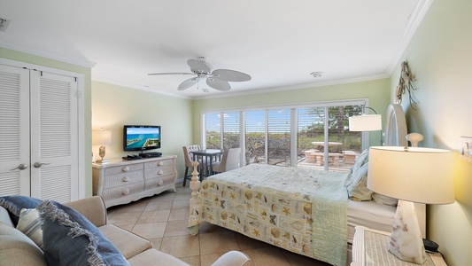 A cozy bedroom with a bed, small sofa, TV on a dresser, and a table with chairs by a large window looking out to an outdoor patio area with greenery beyond.