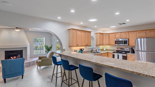 Modern kitchen with granite countertops, stainless steel appliances, and light wood cabinets. A curved archway opens to a sitting area with blue chairs and a lit fireplace.