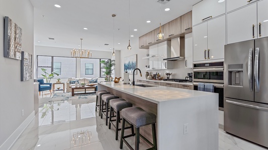 Modern kitchen with a large island and wooden barstools. There are stainless steel appliances, white cabinets, and pendant lights. The kitchen opens into a spacious living area with large windows.