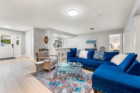 A cozy living room with a blue sectional sofa, a glass coffee table, and a colorful rug. There is a small dining area and a kitchen in the background, with a doorway leading to a bedroom.