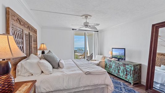 A bedroom featuring a bed with white linens, a ceiling fan, a rustic wooden headboard, a distressed turquoise dresser, a TV, and a sliding glass door leading to a balcony with an ocean view.