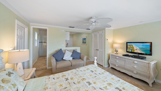 A cozy bedroom features a bed with patterned bedding, a small seating area with a couch, a dresser with a TV displaying a beach scene, and a ceiling fan. A bathroom entry and a framed picture are visible.