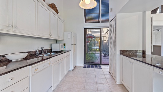 A bright kitchen with white cabinets, a black granite countertop, a dishwasher, microwave, and a white refrigerator. There is a door leading to an outdoor patio area with tables and chairs.