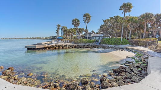 Small secluded beach area with rocky shoreline, clear water, palm trees, and nearby houses on a sunny day.