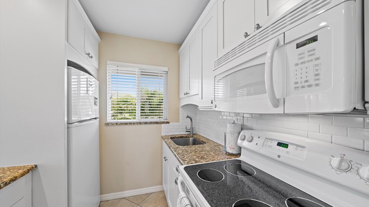 A compact kitchen with white cabinets, granite countertops, an electric stove, microwave, refrigerator, and a small sink beneath a window with blinds.