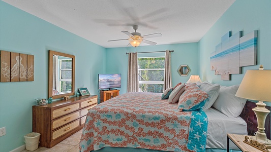 A bedroom with turquoise walls, wood furniture, and a bed with coral-themed bedding. The room has a ceiling fan, a TV on a dresser, a window with gray curtains, and beach-themed decor.