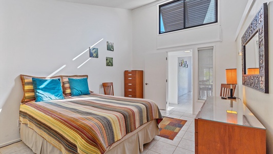 Bedroom with a striped bedspread on a double bed, two blue pillows, wooden furniture, a wall mirror, and a large window near the ceiling. Natural light illuminates the white walls.