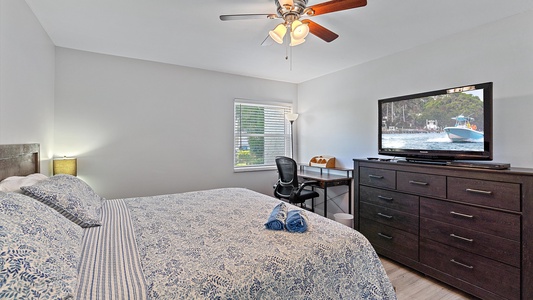 A bedroom with a bed covered in a blue floral-patterned comforter, a desk and office chair, a dresser with a TV showing a boat, a window with blinds, and a ceiling fan with light.