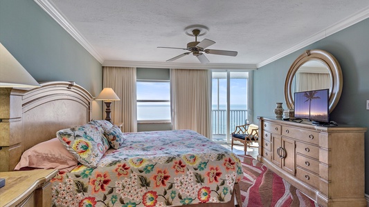 A bedroom with a wooden bed and floral bedding, two lamps, a dresser with a round mirror, a TV, and a balcony offering an ocean view.