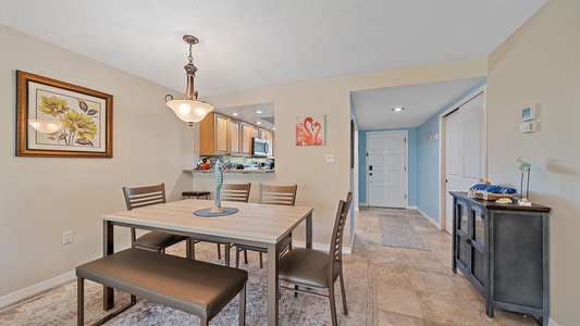 A dining room with a rectangular table and four chairs, a painting on the wall, a pendant light above the table, and a view of the kitchen through an open wall space.