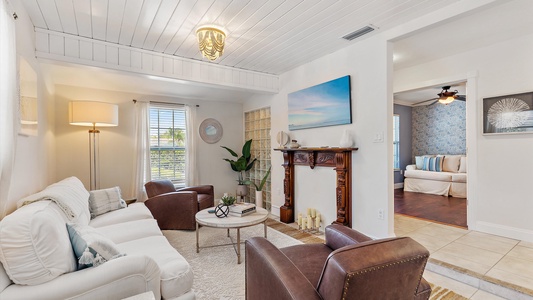 A cozy living room with a white ceiling, white walls, white sofa, brown chairs, a round coffee table, and a decorative fireplace. An adjacent room features a blue patterned wall and a ceiling fan.