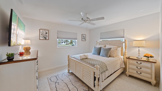 A bright bedroom with a white wooden bed, patterned rug, and off-white dresser. There are two windows, a ceiling fan, a table lamp, plants, and framed artwork on the walls.