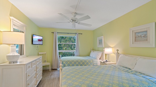 A bedroom with two twin beds, each with a patterned bedspread, a dresser with a lamp, a wall-mounted TV, a ceiling fan, and a window with curtains. Light yellow walls and artwork decorate the room.