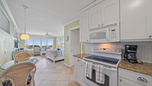 A small, airy apartment with a kitchenette featuring white cabinets, a stove, microwave, and coffee maker. There is a dining table with chairs, a bed, and a seating area by large windows in the background.