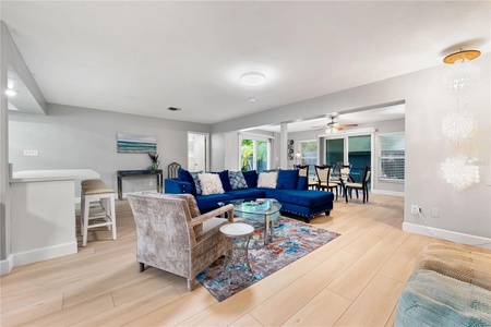 A modern living room with a blue sectional sofa, a gray armchair, a glass coffee table on a colorful rug, a dining area, and large windows with blinds in a well-lit open-concept space.