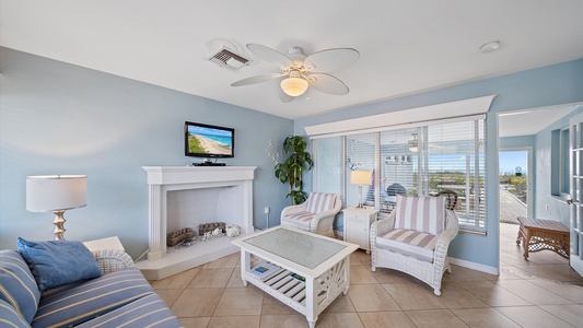 A cozy living room with light blue walls, white furniture, a ceiling fan, a TV above the fireplace, and large windows showcasing an outdoor view. A potted plant is beside the fireplace.