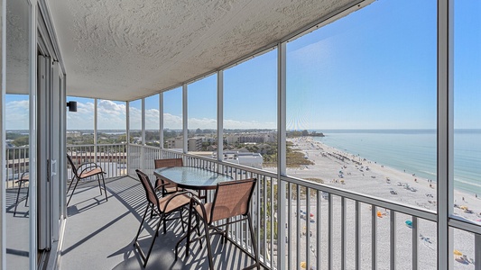 A balcony with a table and chairs offers an expansive ocean view with a sandy beach, clear blue sky, and distant horizon.