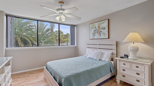 A bedroom with a bed covered in a light blue quilt, a nightstand with a lamp, a dresser, and a ceiling fan. The room has a large window with a view of palm trees and a framed picture on the wall.