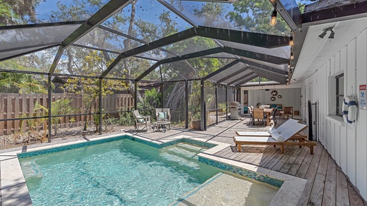 A screened-in swimming pool area with a connected spa, wooden deck, lounge chairs, outdoor dining table with chairs, and surrounding tropical plants. The area has string lights overhead.