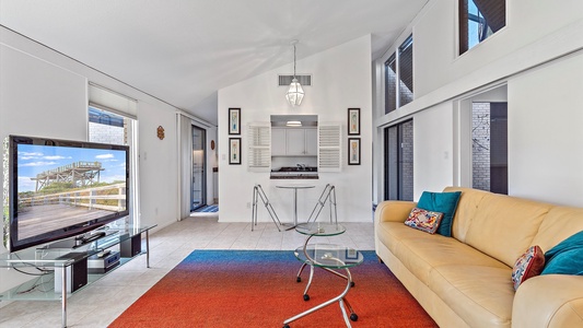 A modern living room with a yellow sofa, glass coffee table, flat-screen TV, and a colorful rug. A small dining area and a kitchen with a pass-through window are visible in the background.