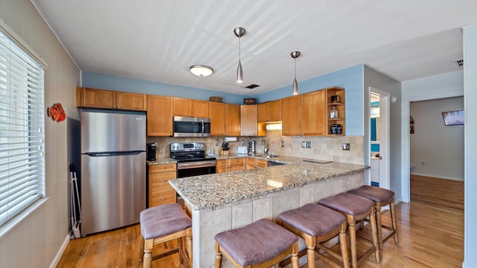 A modern kitchen features wooden cabinets, stainless steel appliances, a large granite island with four stools, hardwood floors, and pendant lighting.